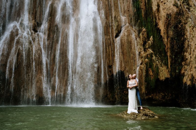 Bride and groom standing on rock in the water beneath Limon Waterfall in Las terrenas