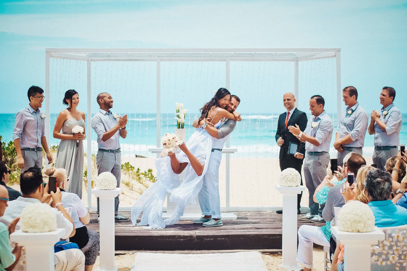 Bride and groom embracing at their beach altar as the wedding party celebrates around them at a Punta Cana destination wedding