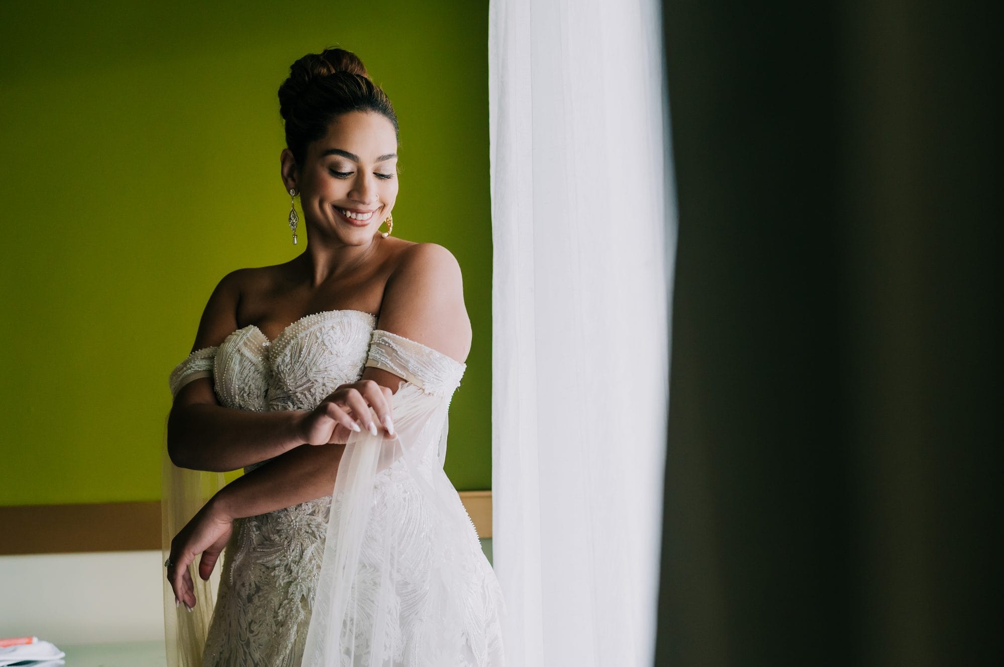 Bride adjusting the off-the-shoulder sleeves of her beaded lace wedding gown by the window