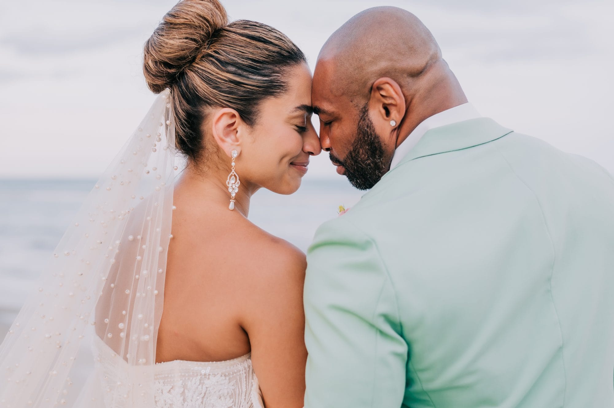Bride and groom forehead to forehead by the ocean, pearl veil flowing, at Dreams Onyx Punta Cana