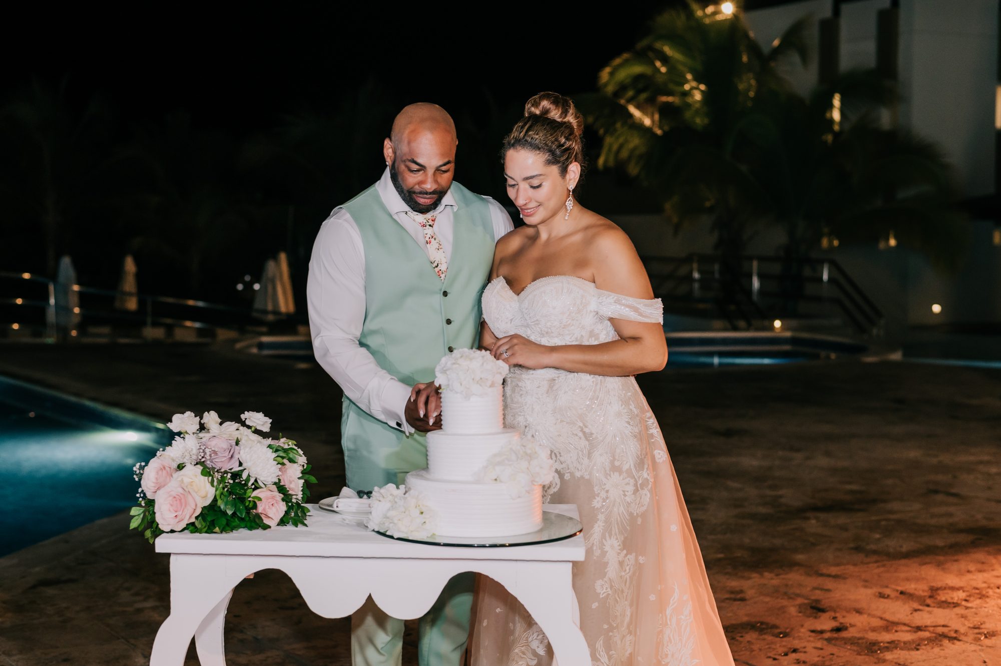 Bride and groom cutting their three-tier floral wedding cake poolside at night at Dreams Onyx