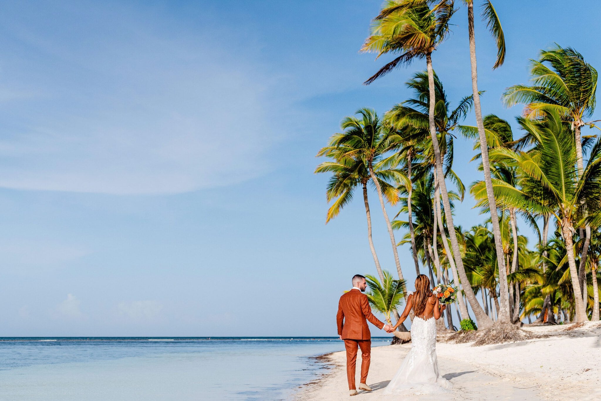 Bride and groom walking hand in hand along a sunny palm-lined Punta Cana beach