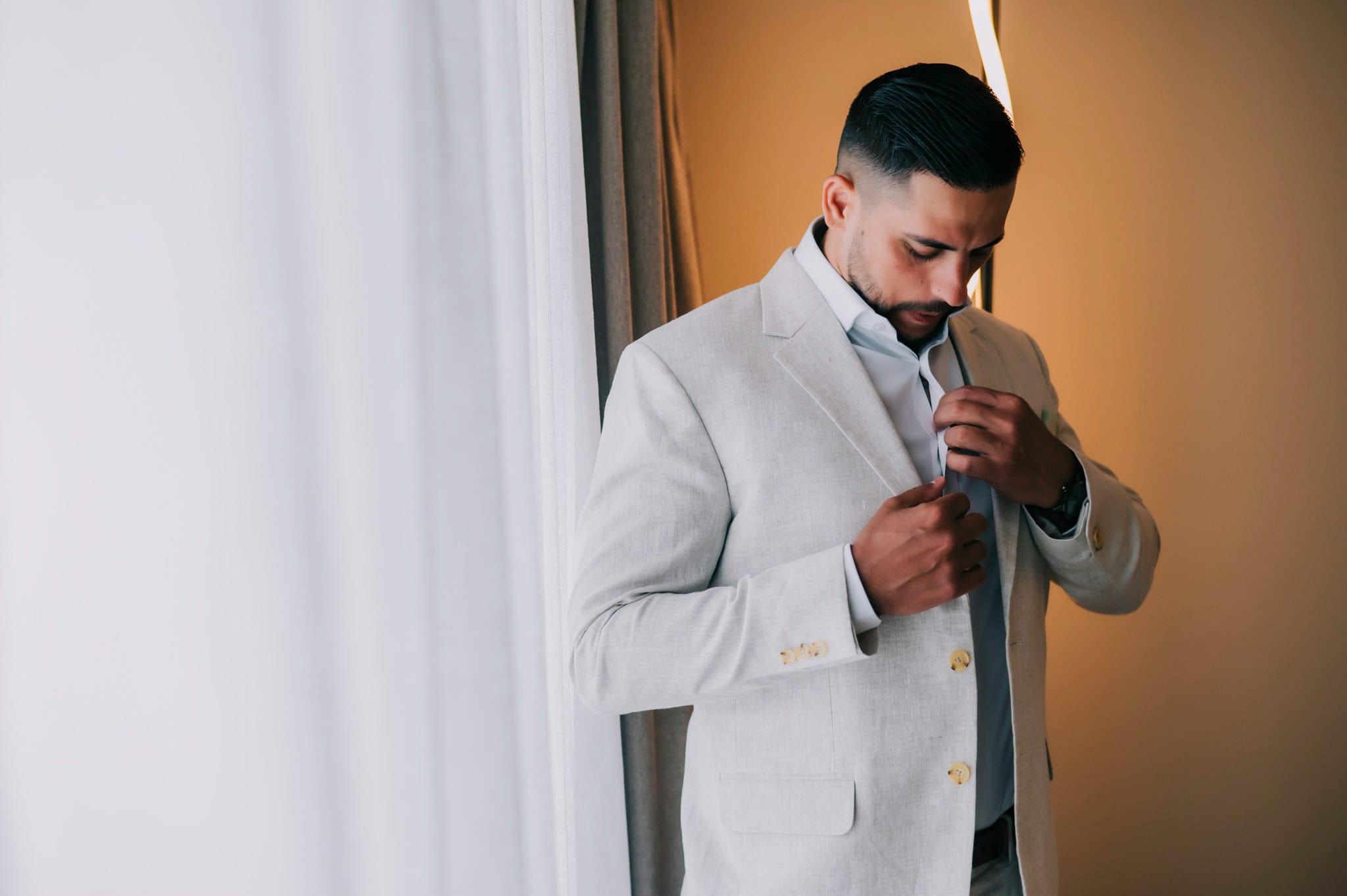 Groom adjusting his jacket and tie beside a window while getting ready for the wedding.