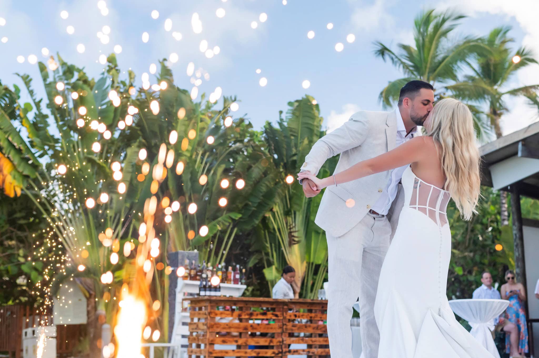 Wide first-dance view of the newlyweds framed by spark fountains and tropical greenery.