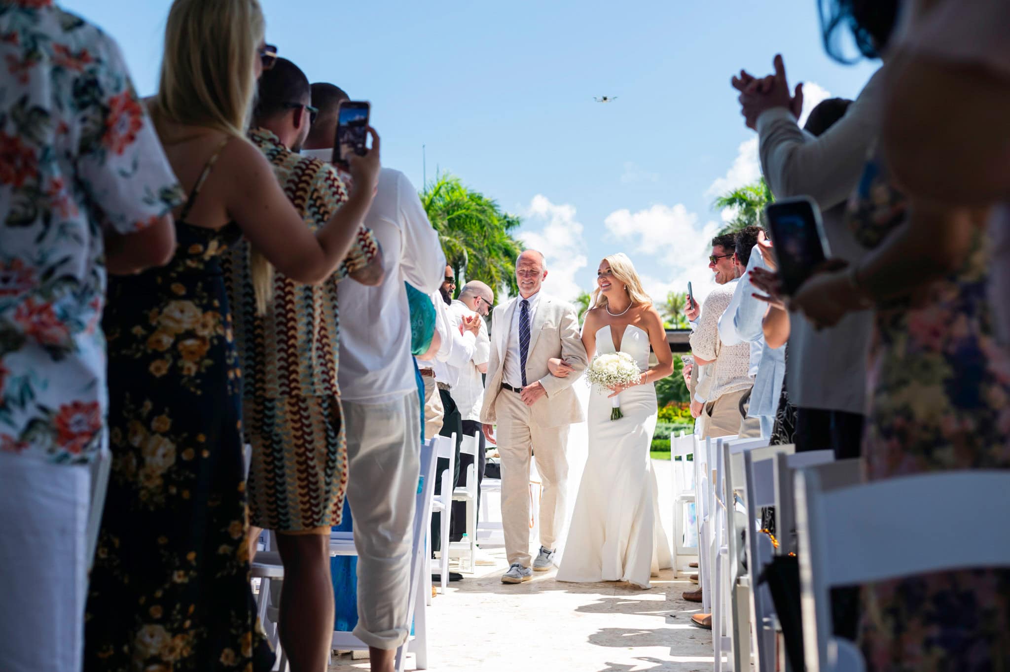 Bride walking down the resort aisle with her father as guests applaud and record the processional.
