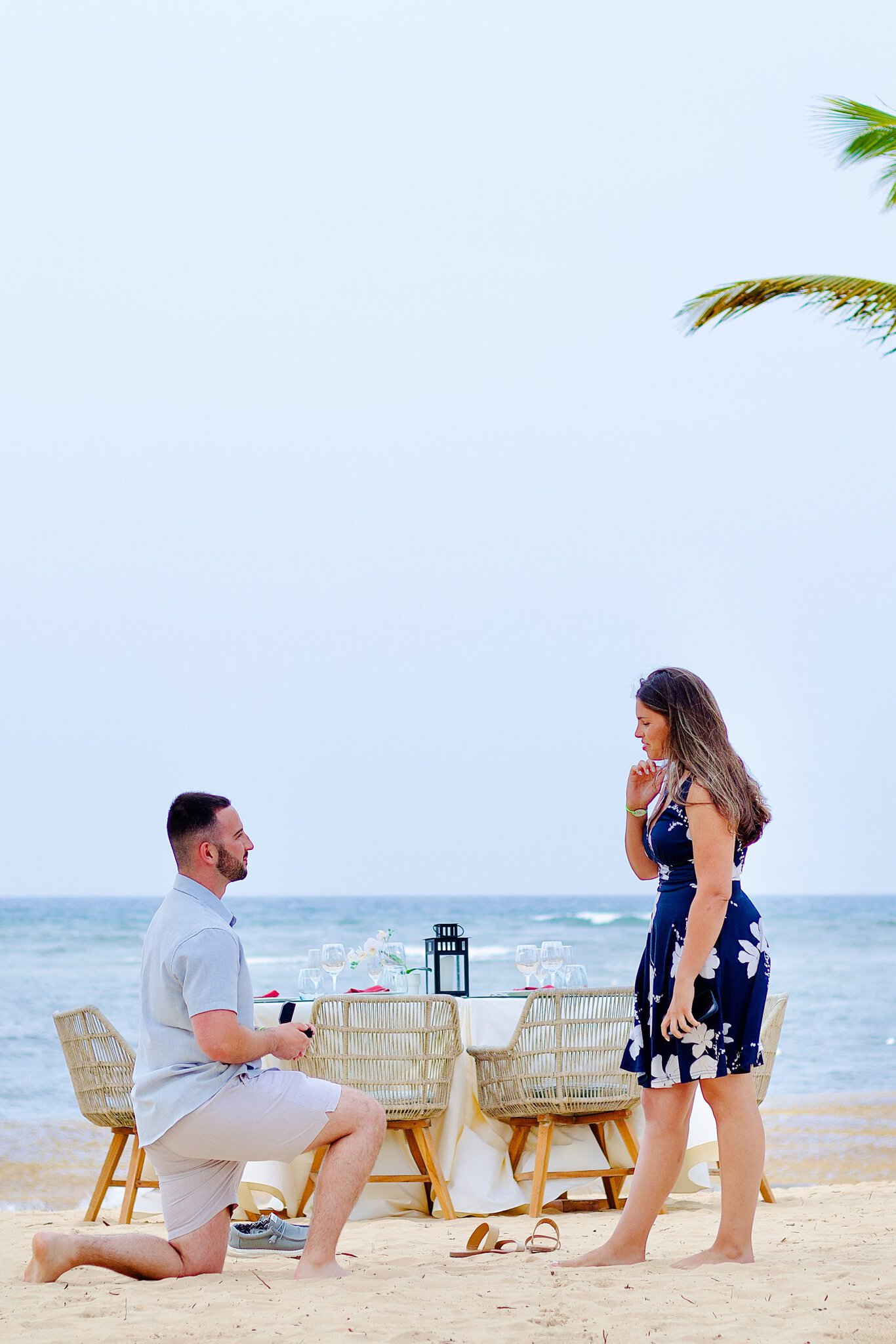 Man kneeling on the beach at Live Aqua Punta Cana to propose at sunset as his surprised girlfriend stands by the decorated dinner table