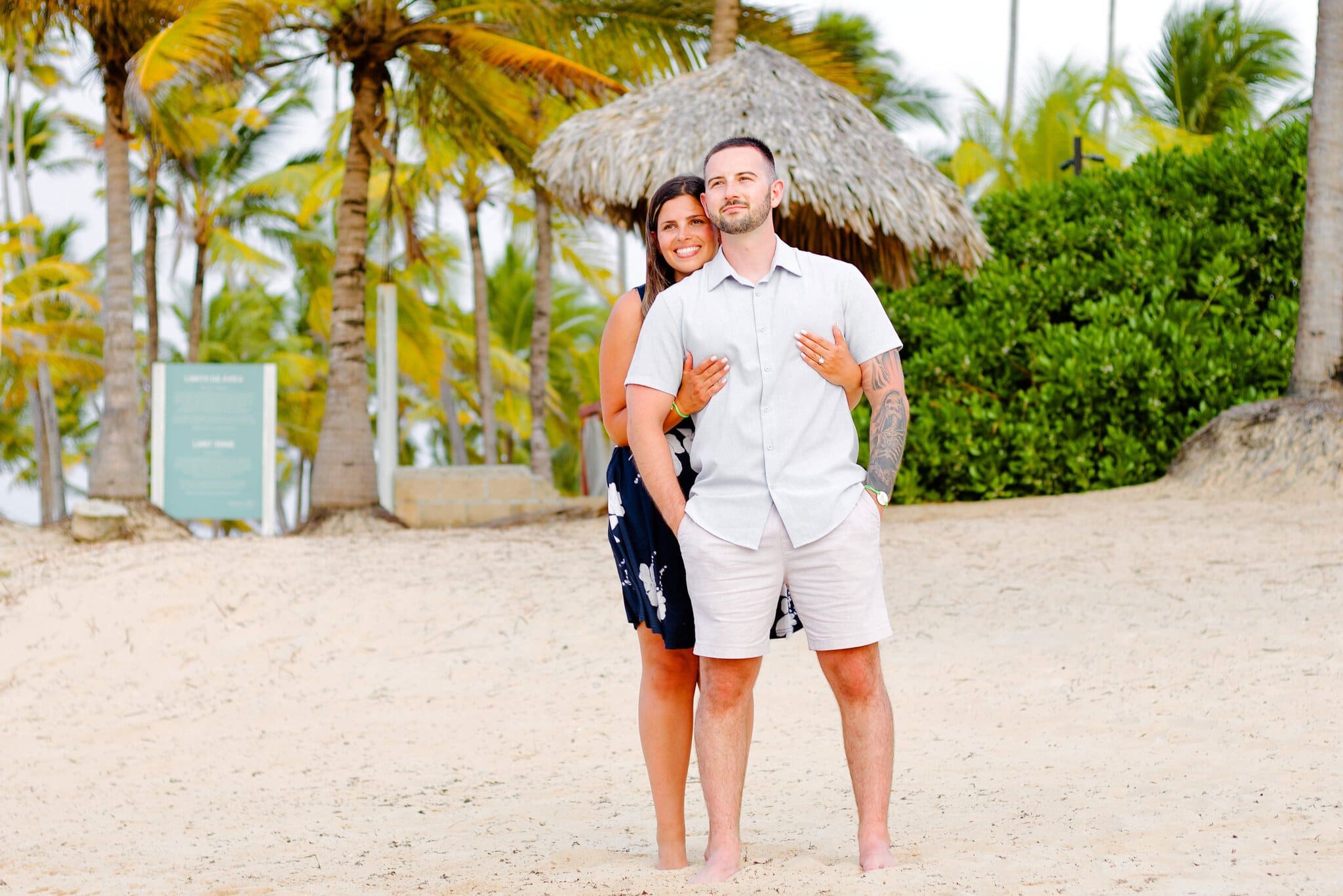 Engaged couple embracing near a thatched palapa and palm trees on the beach