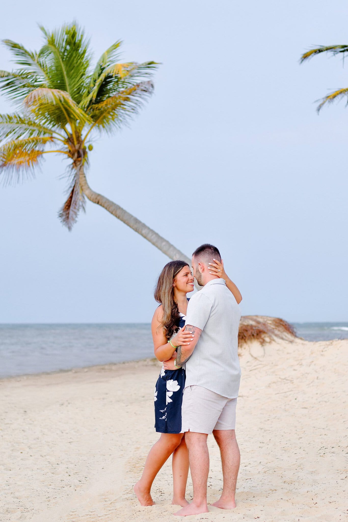 Engaged couple forehead to forehead under a palm tree with the Caribbean behind them