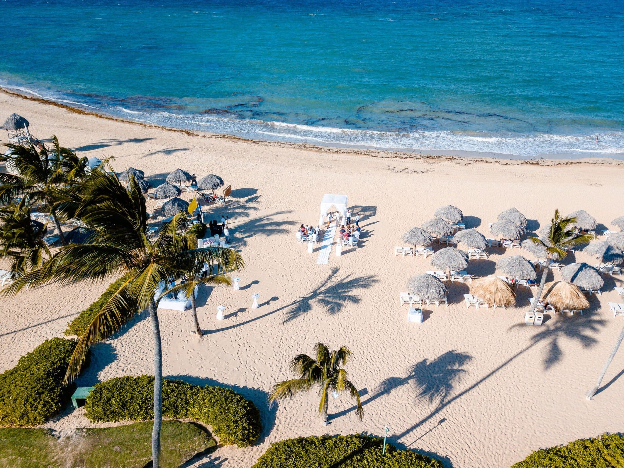 Aerial view of a small beach wedding ceremony in Bávaro with palm trees, palapa umbrellas and turquoise ocean water