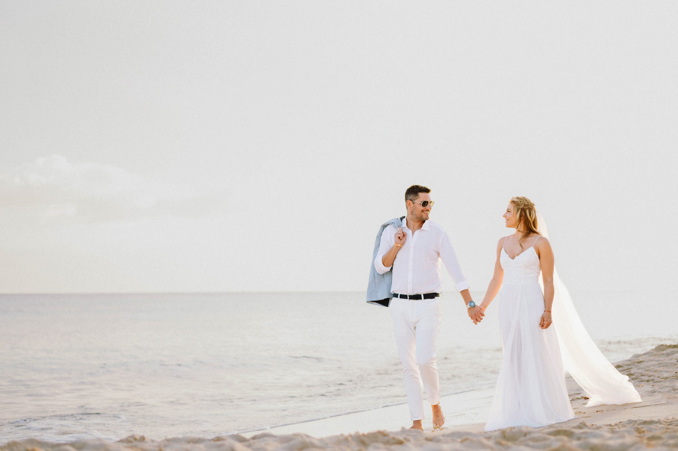 Bride and groom walking barefoot hand in hand along the shoreline in Bayahibe, the groom carrying his jacket over his shoulder