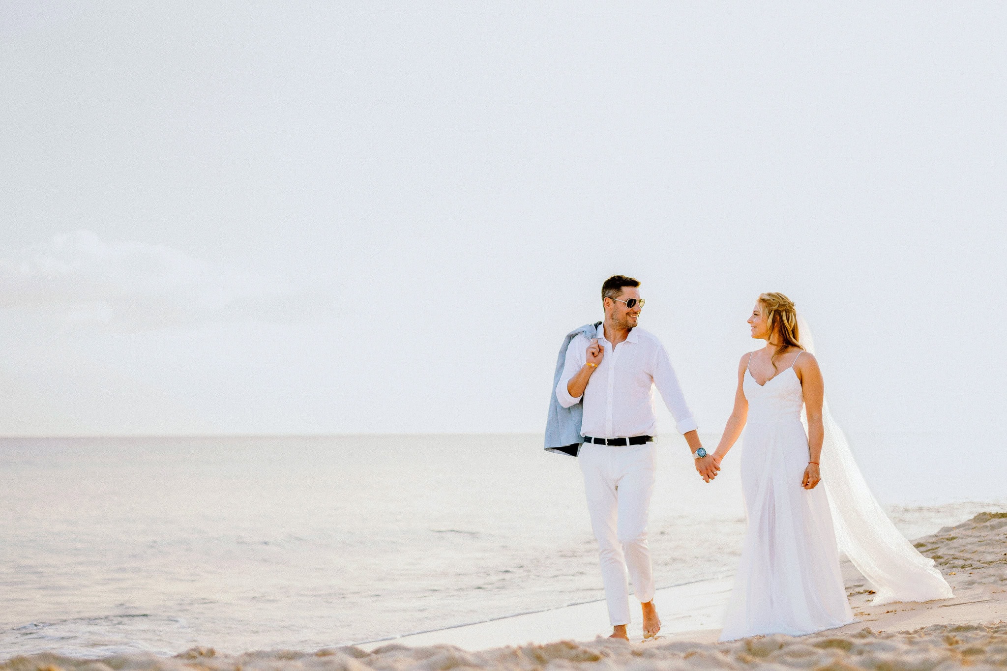 Bride and groom dressed in white walking hand in hand along the shoreline in Bayahibe, Dominican Republic