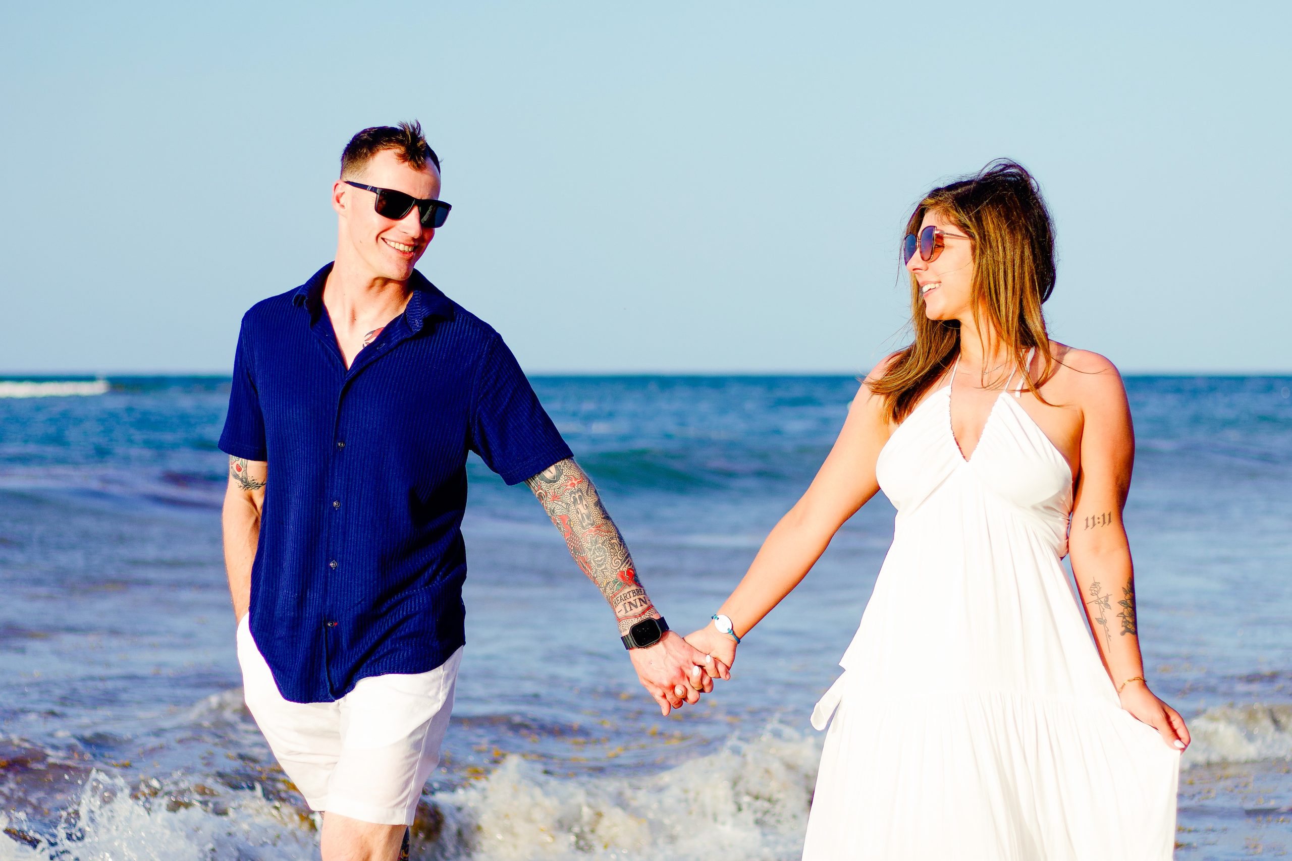 Couple walking hand in hand through the shallow water, smiling at each other in sunglasses and light beachwear, with the open sea behind them