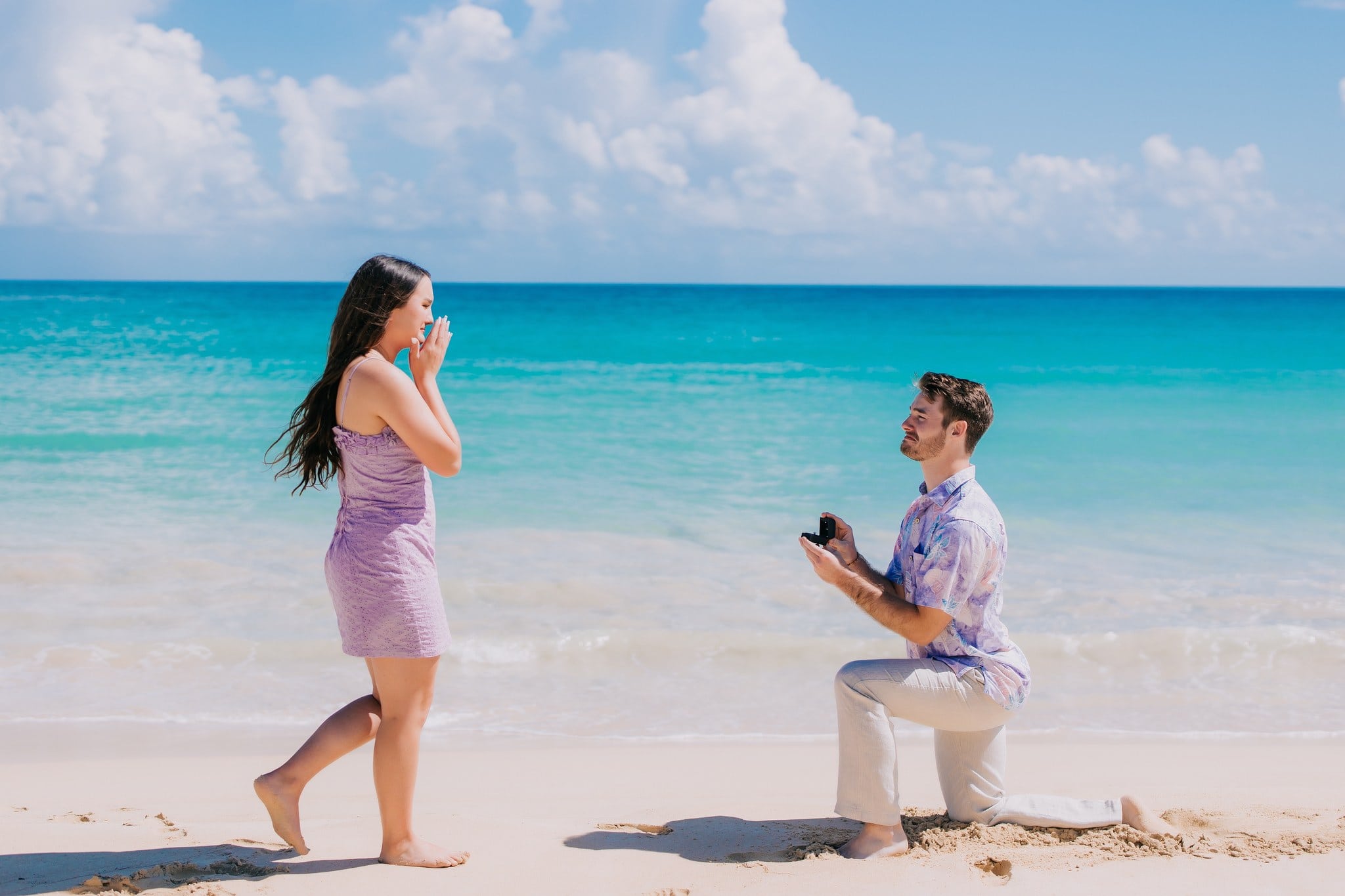 Man on one knee holding an open ring box as his partner brings her hands to her face on a white sand beach