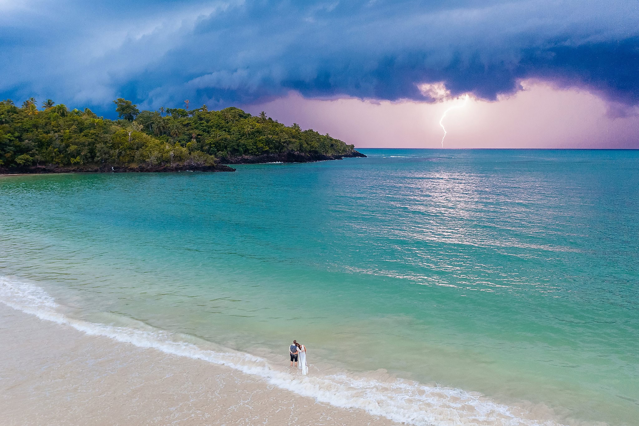 Bride and groom alone at the water edge of a green cove on the Samana peninsula, with lightning breaking over the sea behind them, Dominican Republic