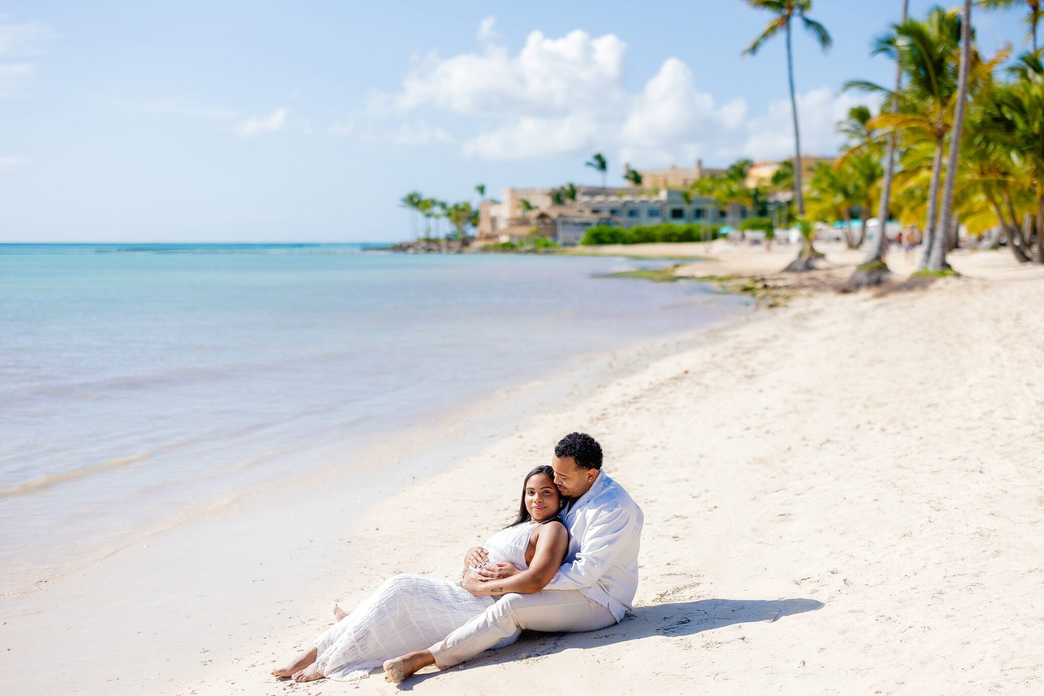 Couple sitting together on a white-sand beach with calm turquoise water and palm trees in Cap Cana
