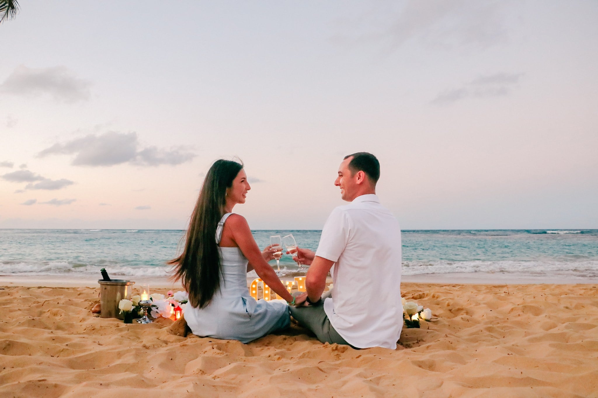 Newly engaged couple sitting on the sand toasting with champagne beside candles and lit LOVE letters