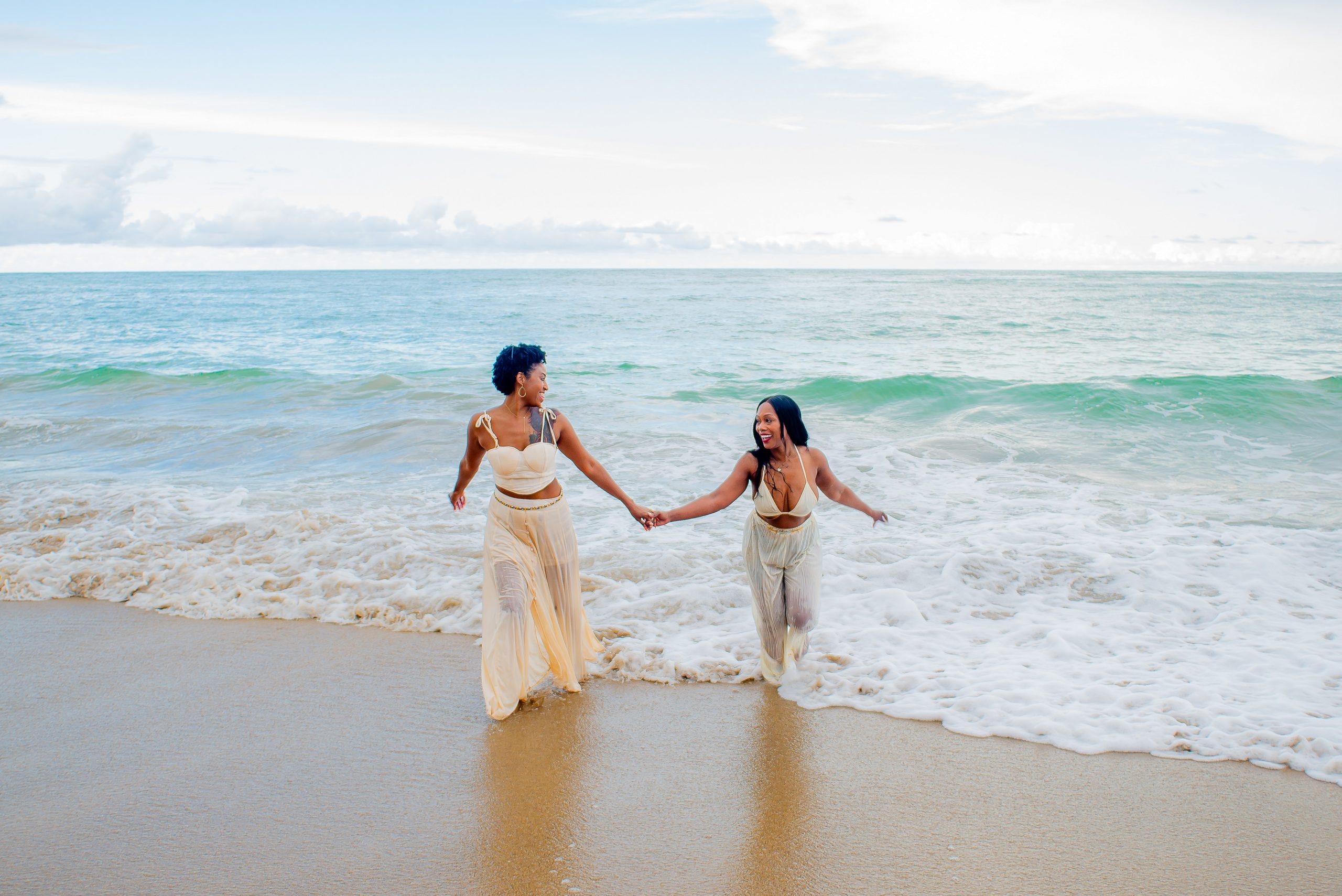 Couple holding hands and laughing together as waves wash over the shore around their feet
