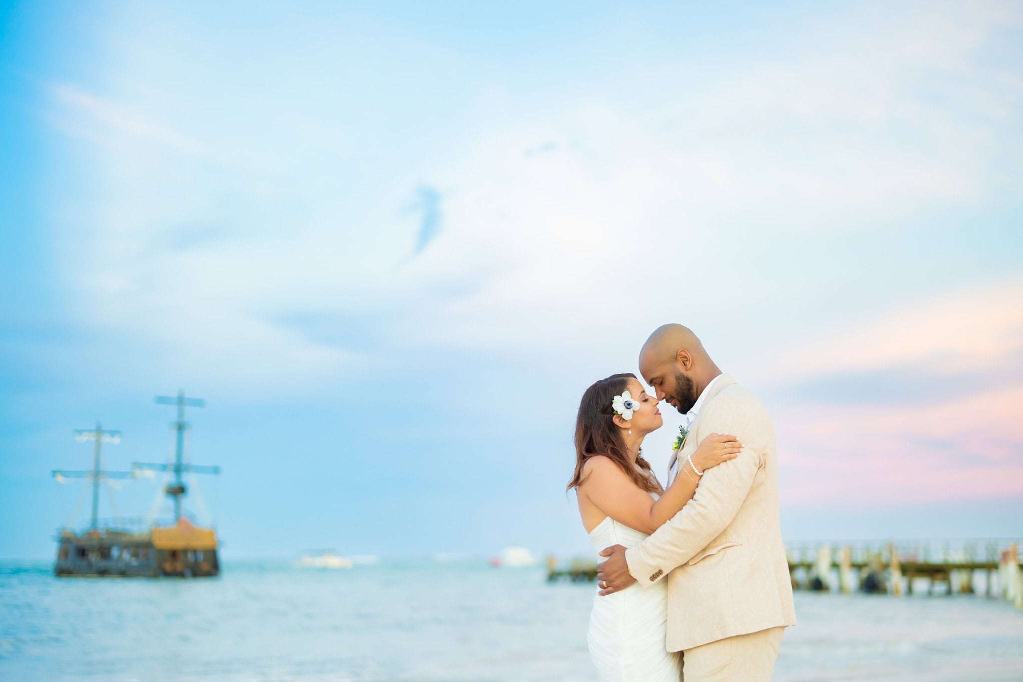 Bride and groom forehead to forehead on the beach at dusk during their Punta Cana elopement, with a tall ship and a pier behind them