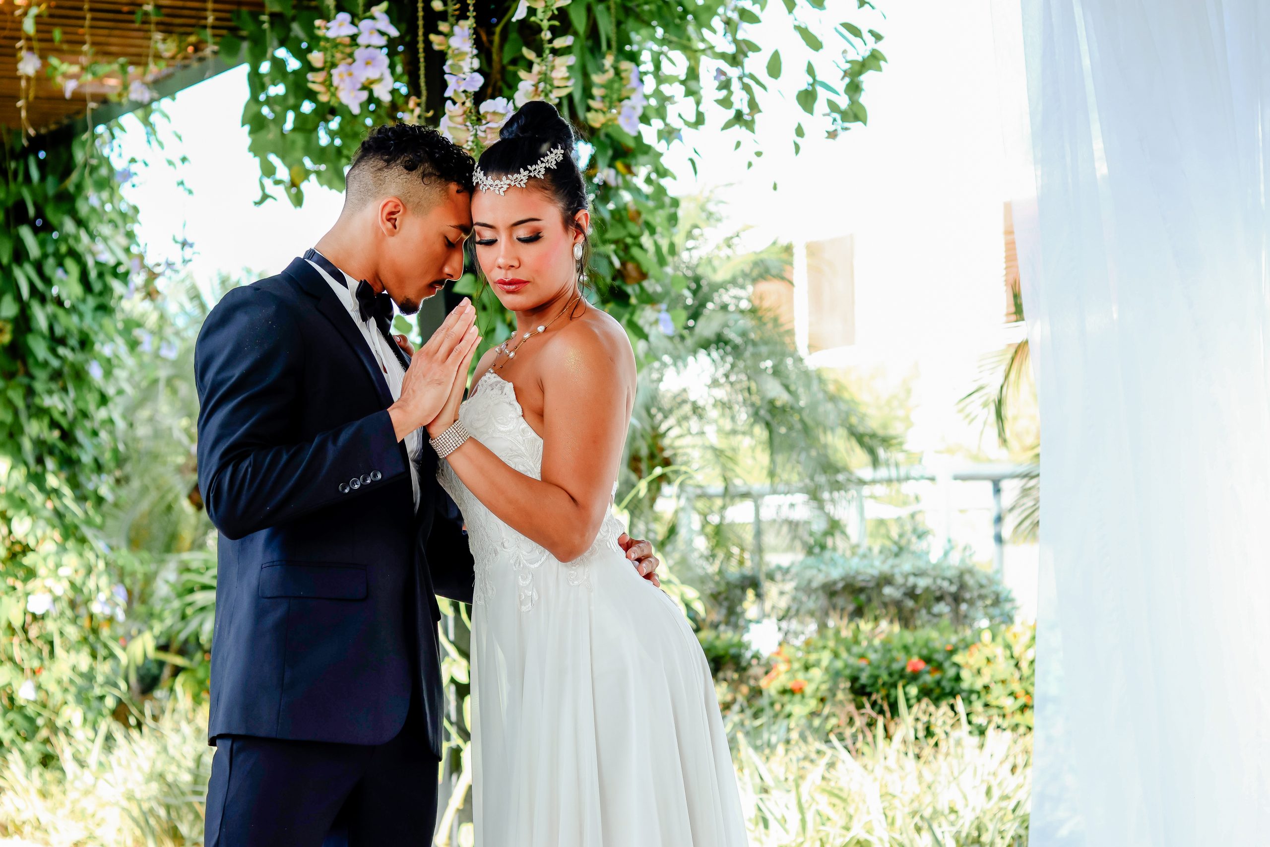 Elopement couple leaning close with their hands pressed together beneath hanging flowers and tropical greenery