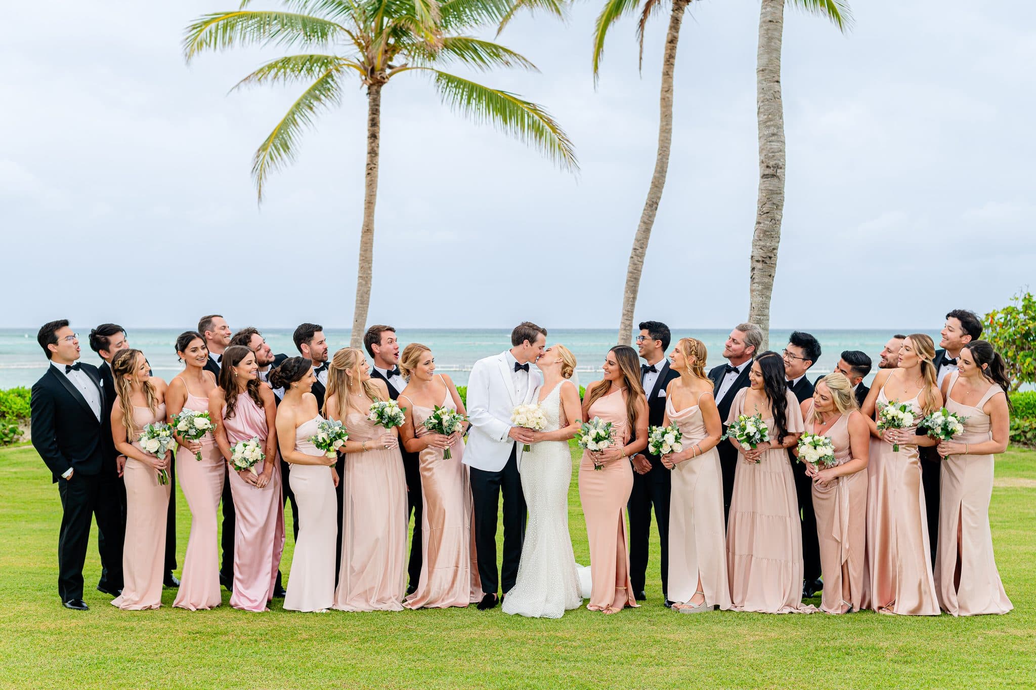 Full wedding party gathered around the kissing couple beneath Punta Cana palm trees