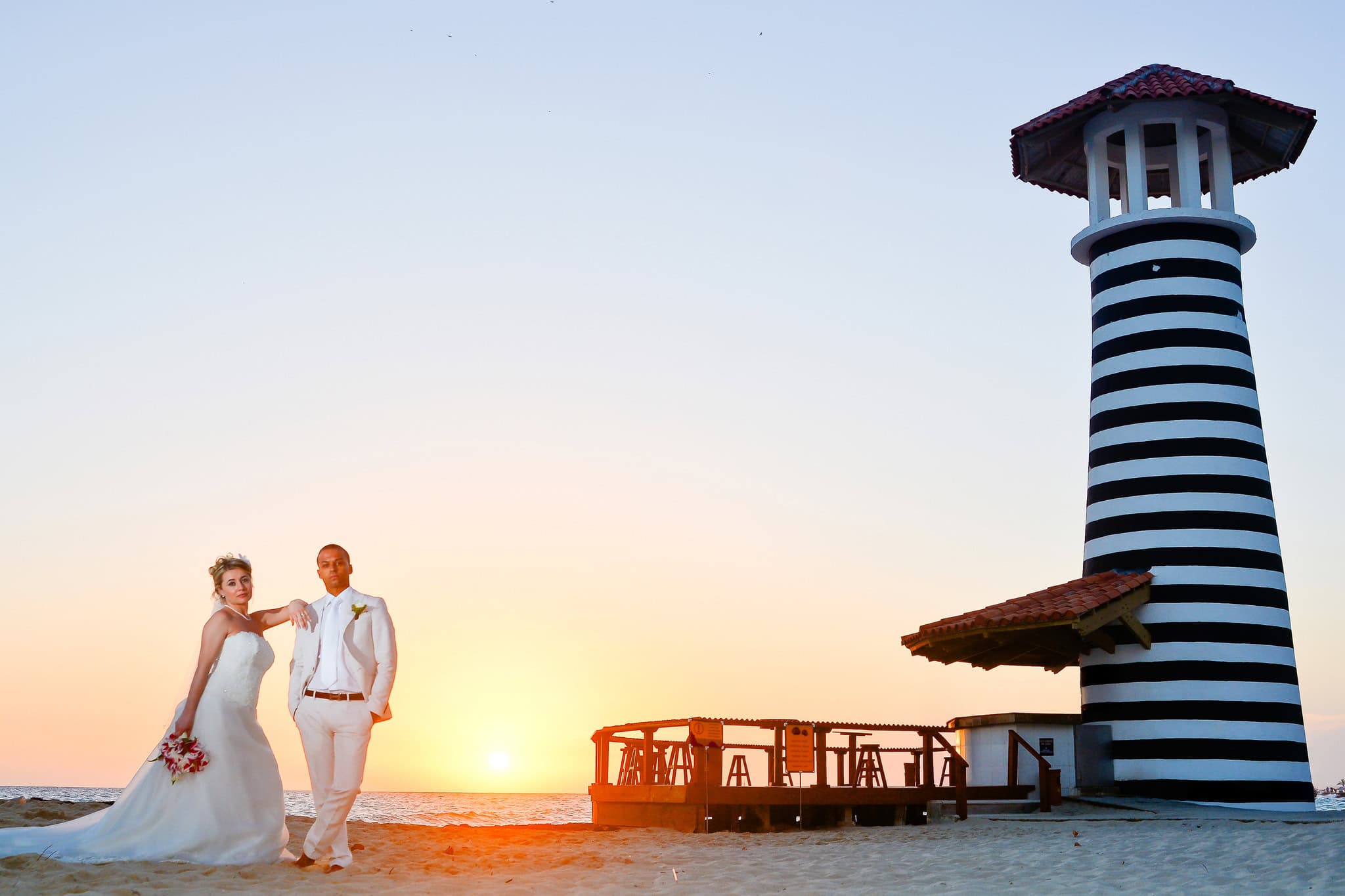 Bride and groom walking on the beach at sunset beside the striped lighthouse at Iberostar Hacienda Dominicus in Bayahibe, Dominican Republic