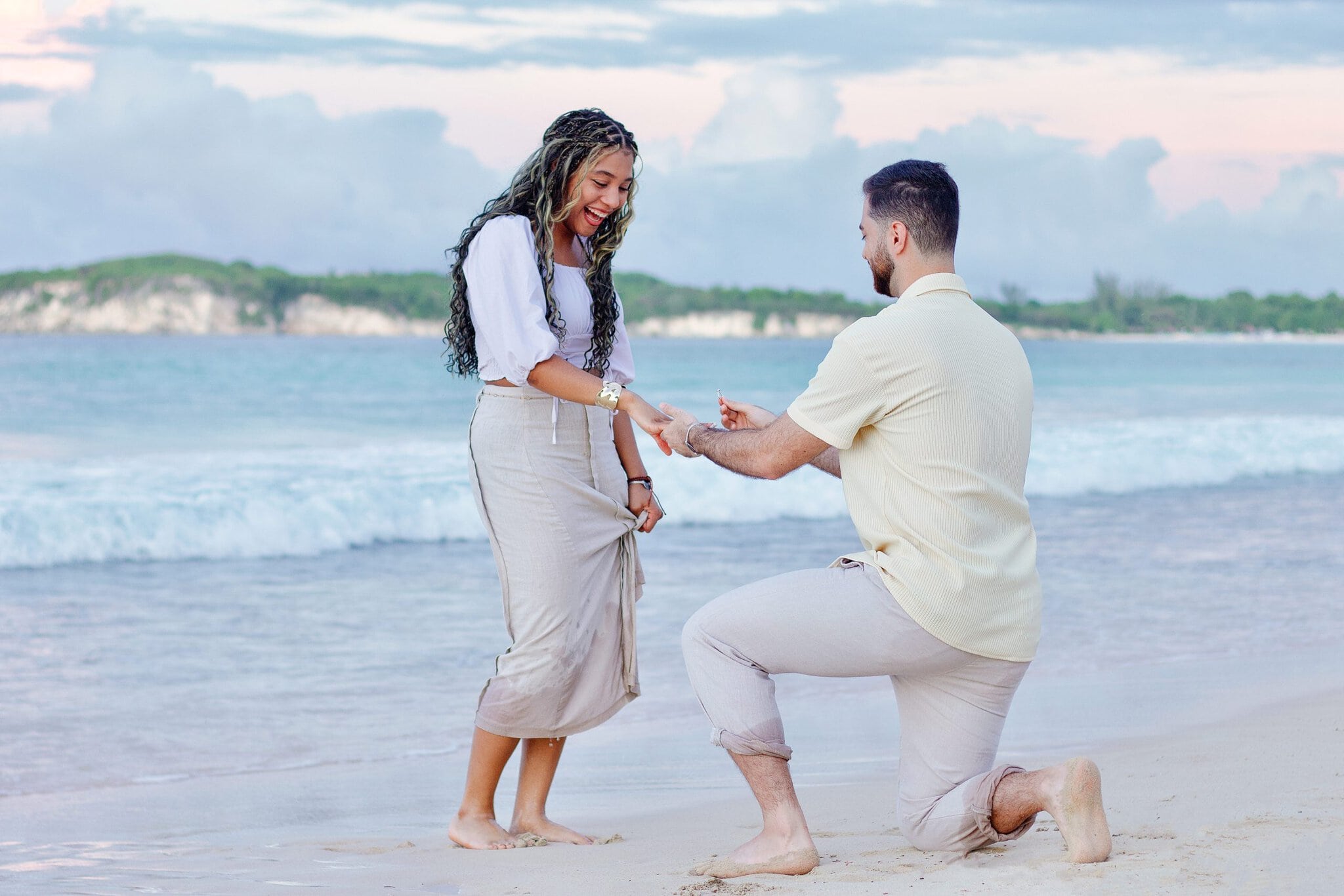 Woman laughing and reaching for her partner’s hand as he proposes with a ring beside the ocean