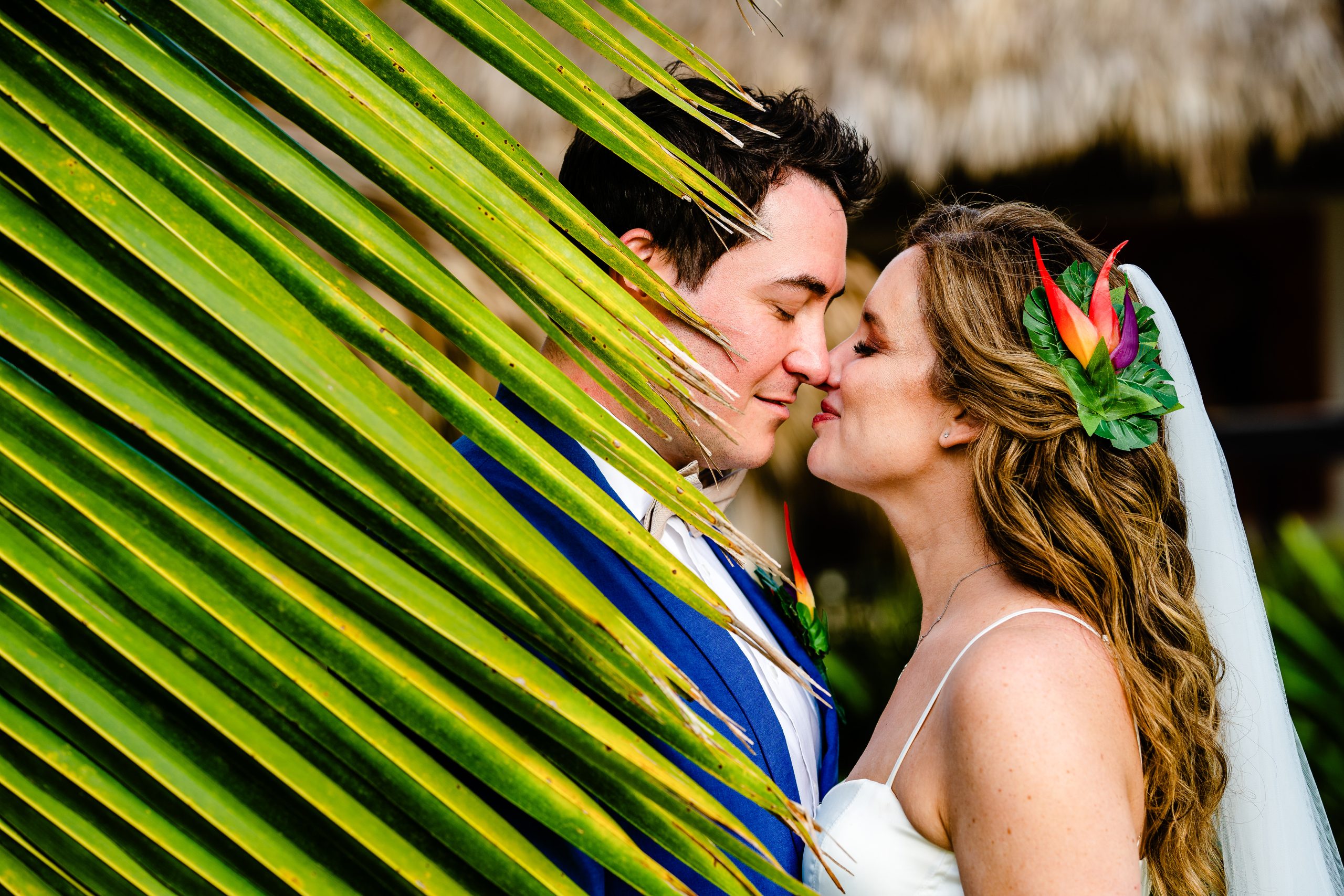 Bride with a tropical flower in her hair and groom in a blue suit about to kiss behind a sunlit palm frond at Los Nómadas in Las Terrenas
