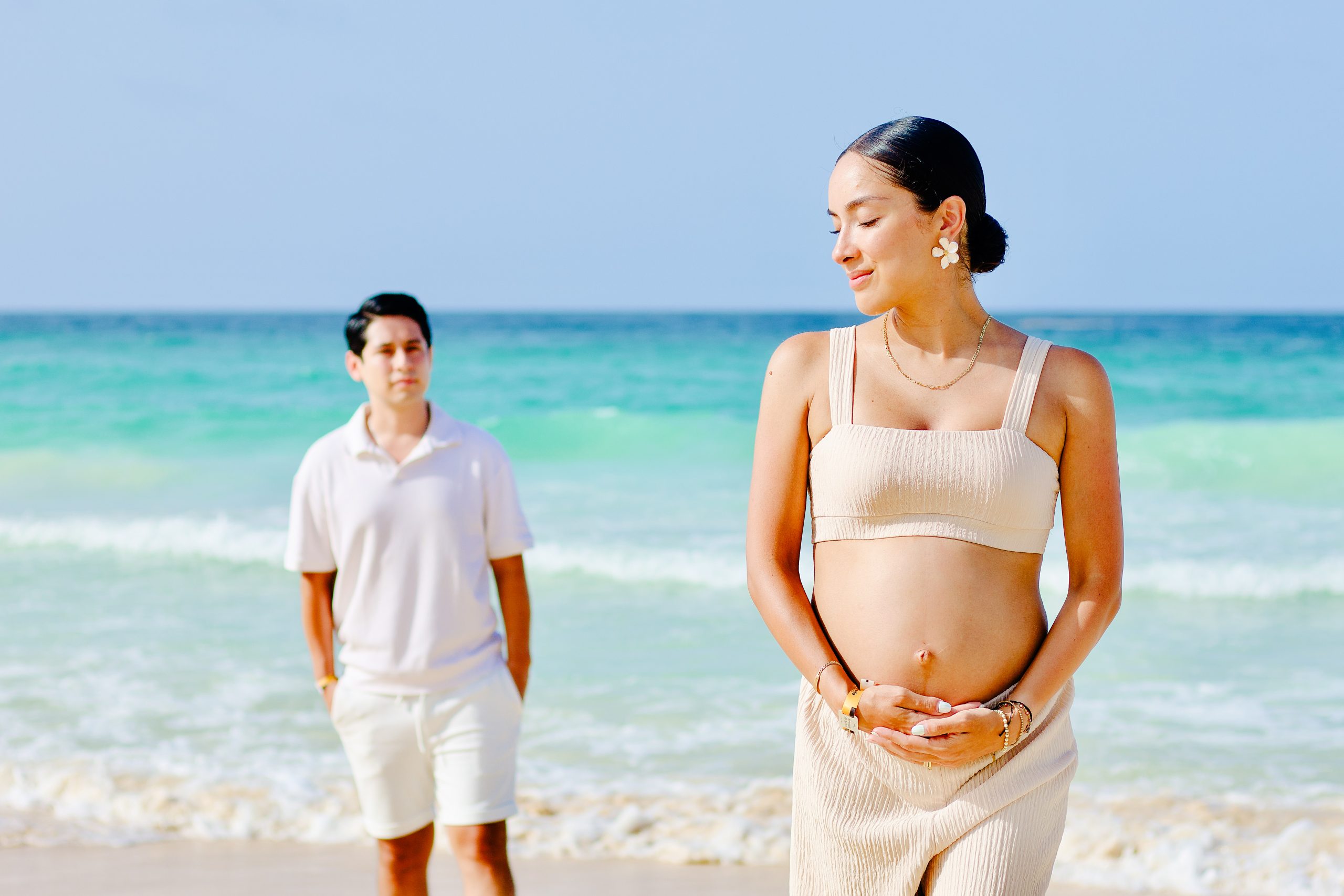 Expectant mother in a cream two-piece resting both hands on her bump while her partner stands behind her on the sand, turquoise sea in the background