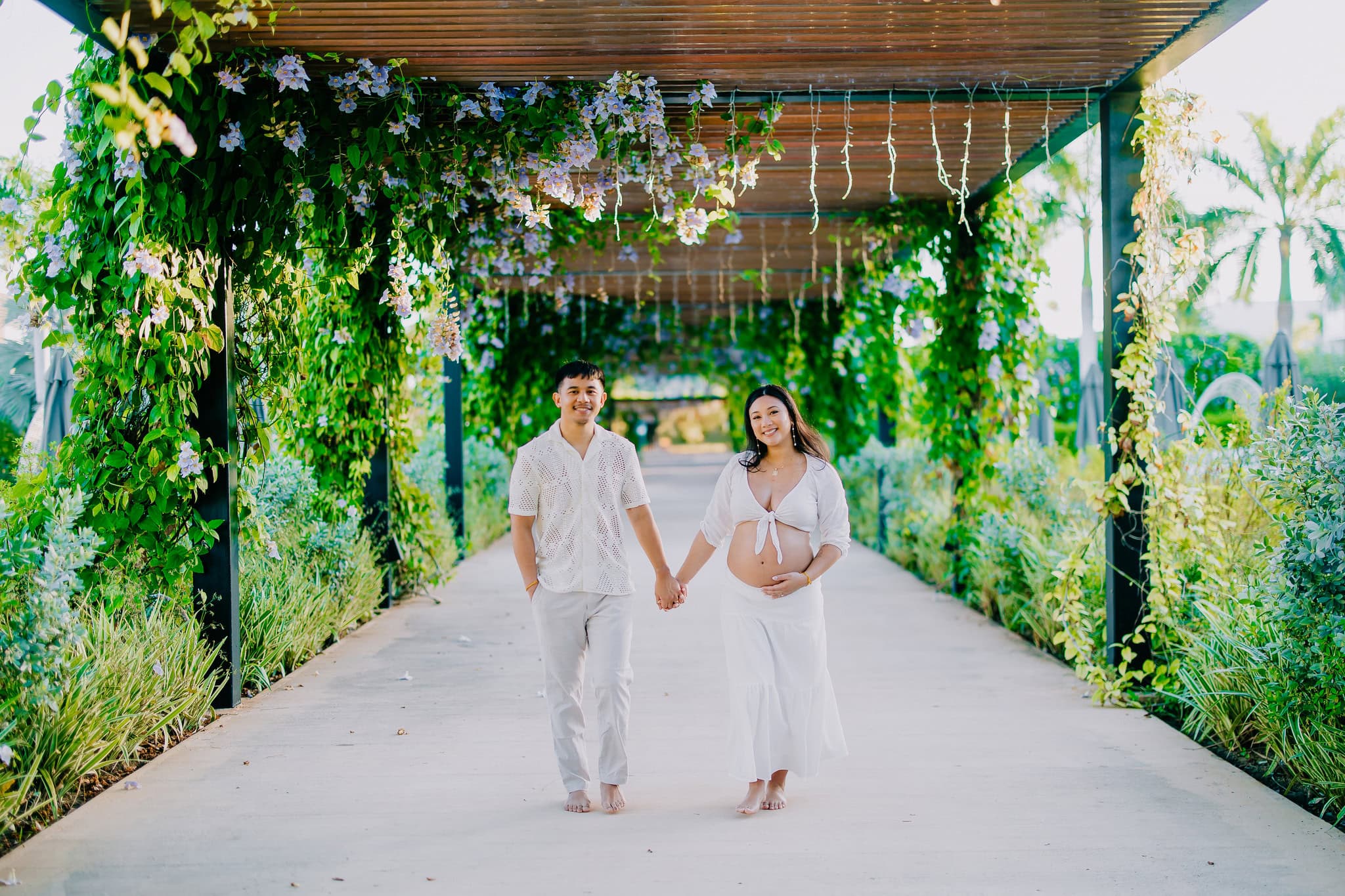 Expecting couple walking hand in hand under a flowered pergola during a maternity session at Live Aqua in Punta Cana