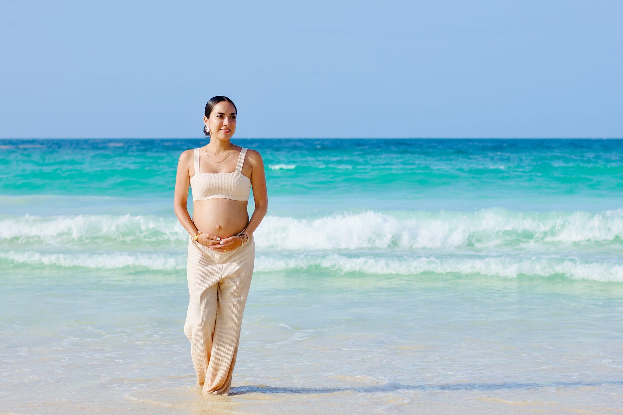Expectant mother standing in shallow ocean water with her hands resting beneath her baby bump