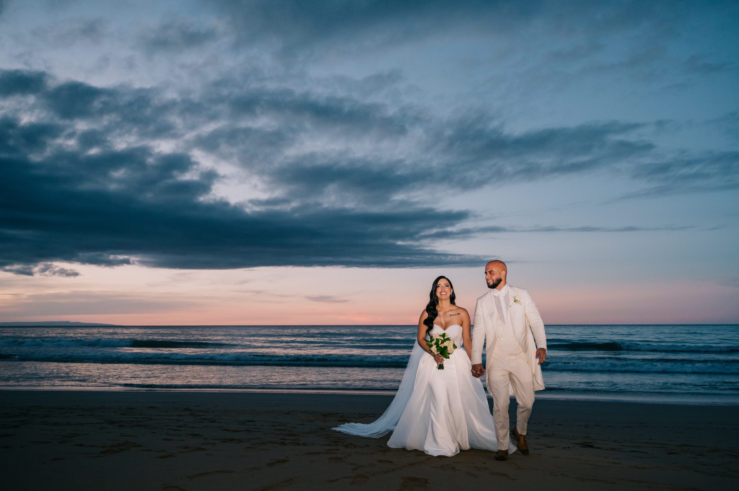 Bride and groom dressed in white walking hand in hand on Playa Cosón beneath dramatic clouds at twilight