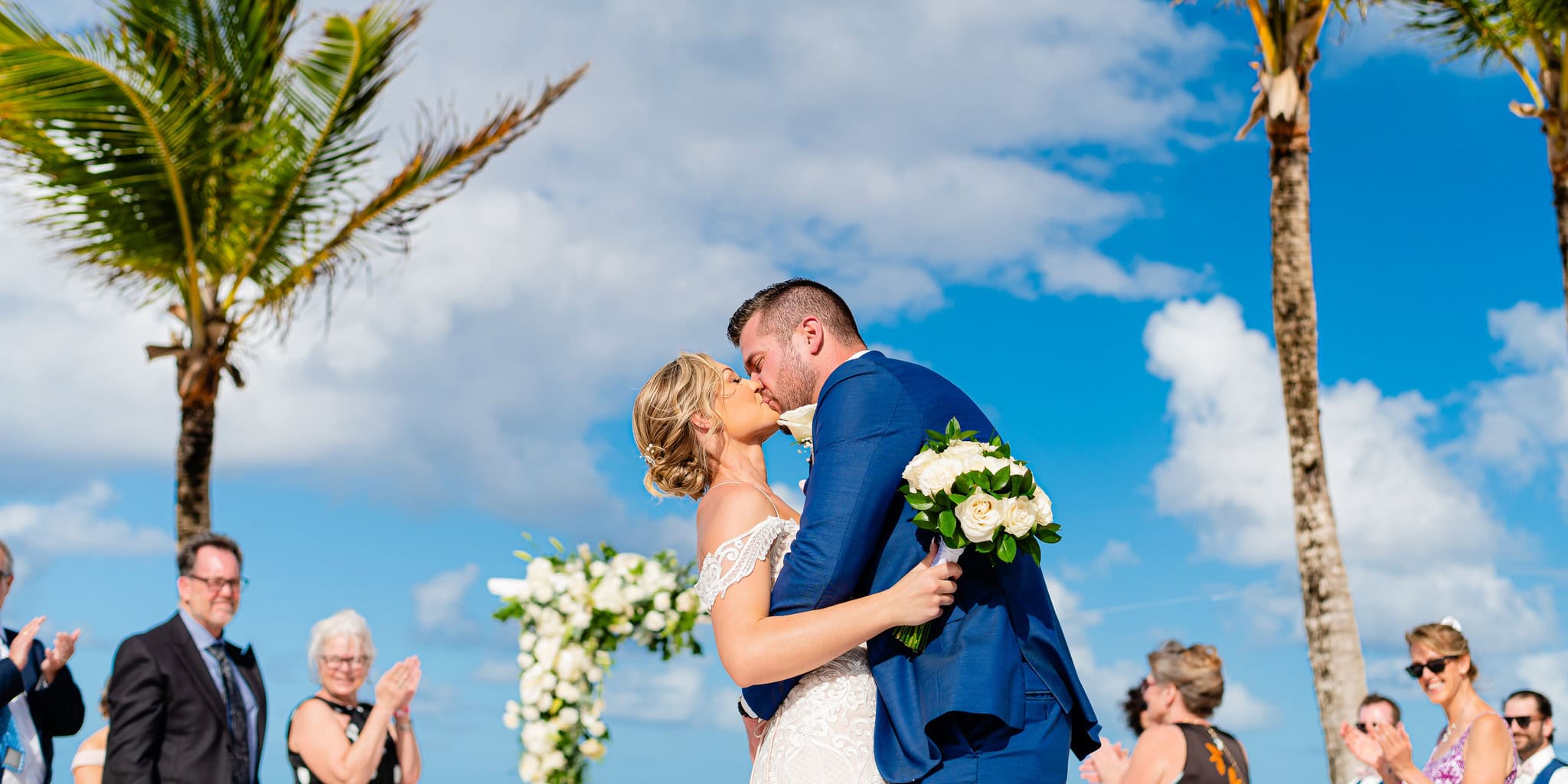 Bride and groom share their first kiss under palm trees as guests applaud at a beach wedding in Punta Cana