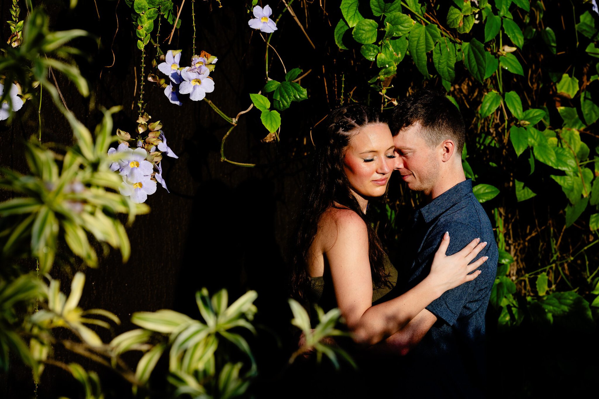Couple embracing beside dense green vines, broad tropical leaves and hanging purple flowers during a photo session in Punta Cana