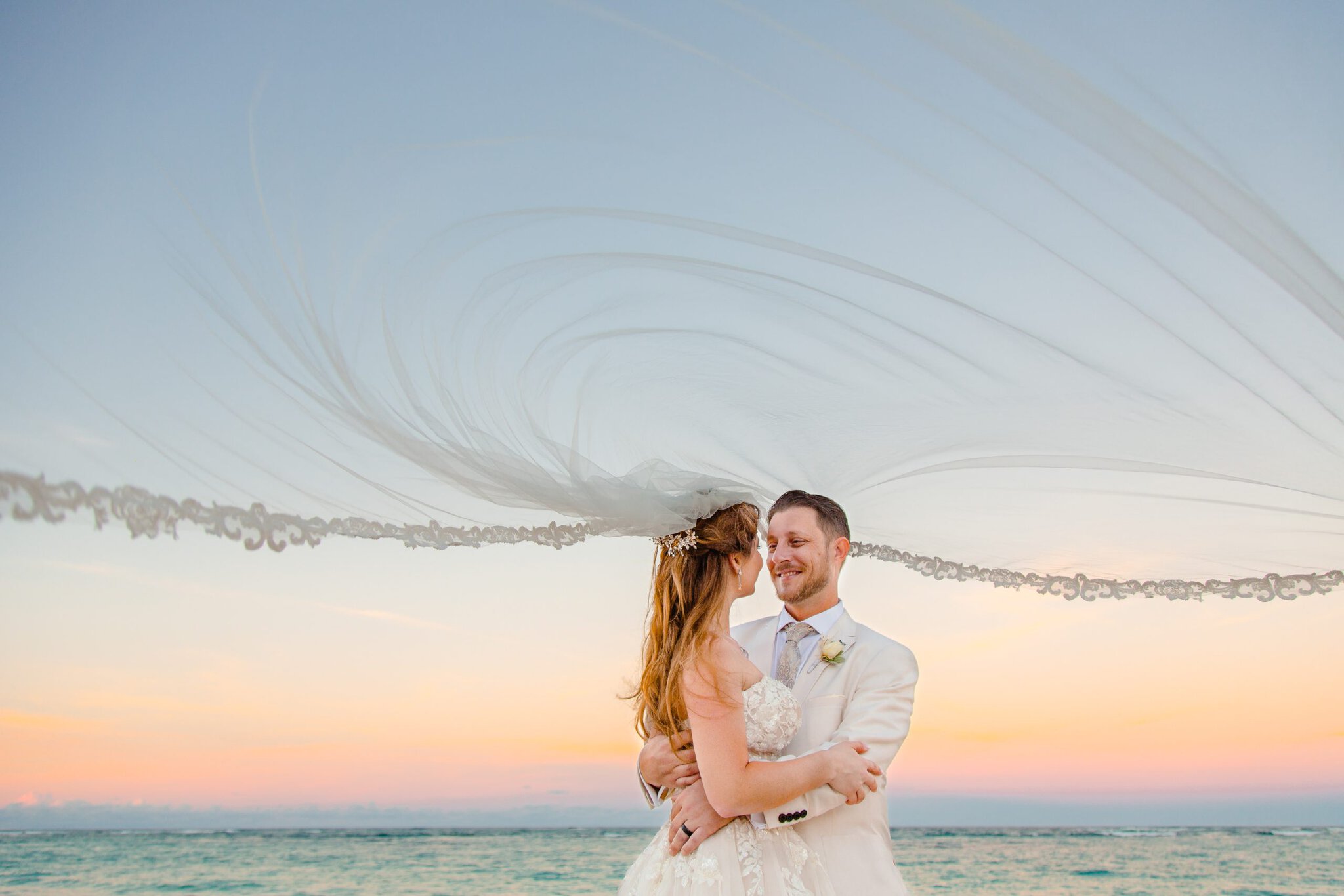 Bride and groom embracing beneath a flowing veil during a sunset wedding portrait in Punta Cana