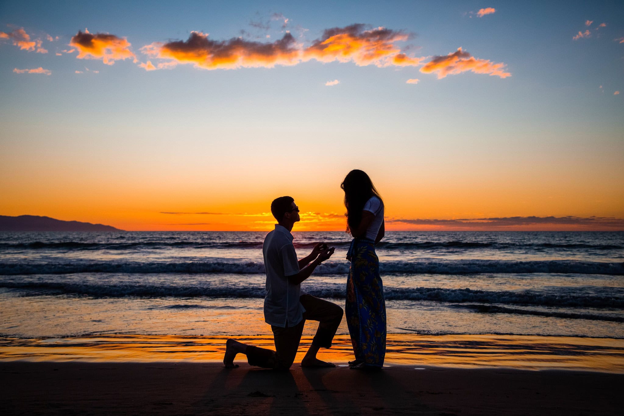 Silhouette of a couple against an orange sky as he proposes on one knee at the water's edge