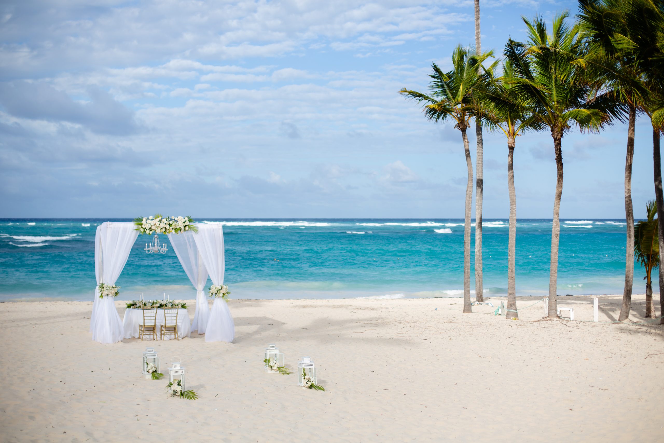 Simple beach ceremony setup with a white canopy, floral details, and palm trees beside the ocean