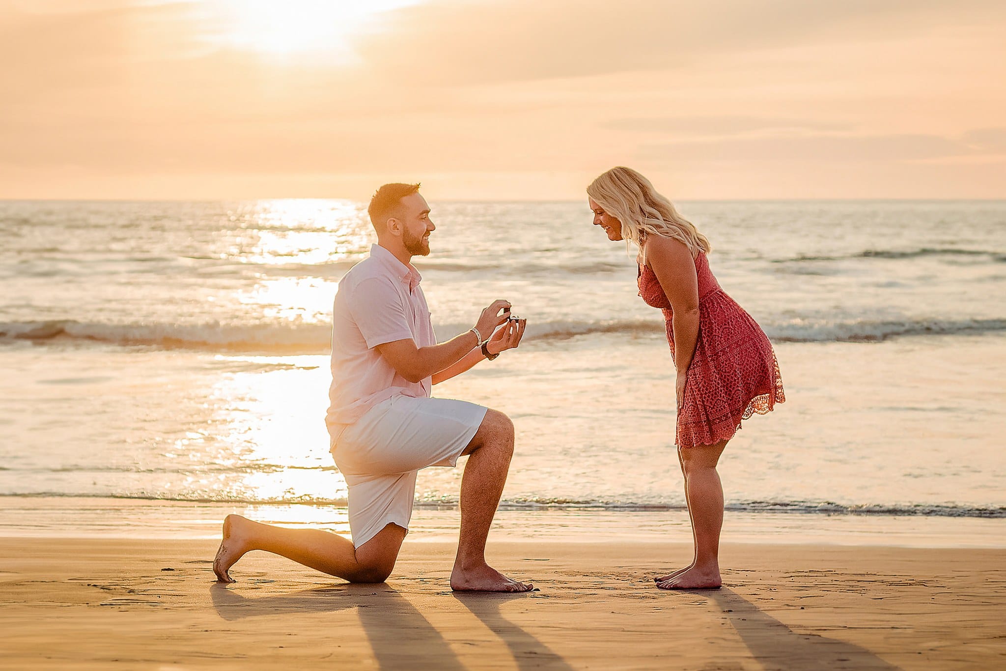 Man proposing on one knee to his partner on the beach as the sun sets behind them