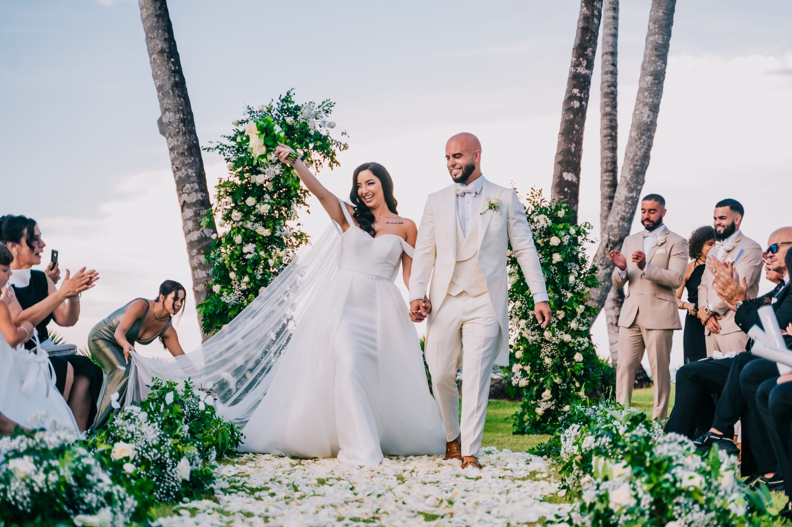 Bride raising her bouquet while walking hand in hand with the groom down a flower-covered aisle at Villa La Cassona in Las Terrenas