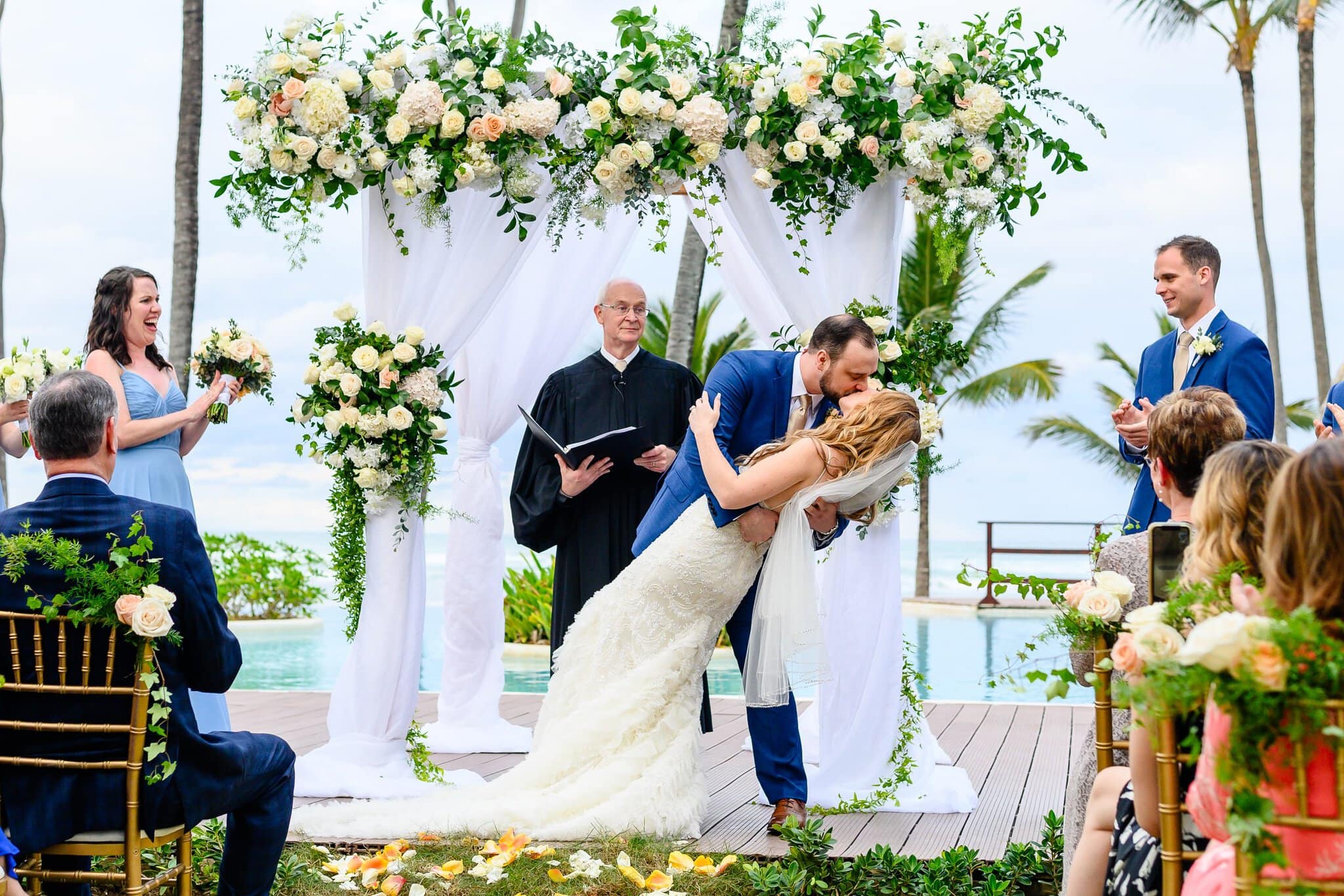 Bride and groom sharing a dip kiss beneath a floral arch as their wedding party and guests celebrate